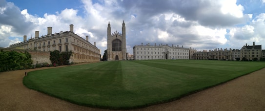 A large open grassy courtyard is bordered by historic stone buildings with numerous windows and chimneys. In the center, a prominent gothic-style chapel stands with intricate architecture and tall spires. The sky is partly cloudy, creating a dramatic backdrop for the scene.