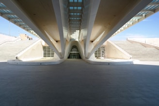 A modern architectural structure with prominent support beams and a symmetrical design. The building is framed by large staircases on either side, leading to an open entrance in the center. Natural light illuminates the area from above, highlighting clean lines and geometric shapes.