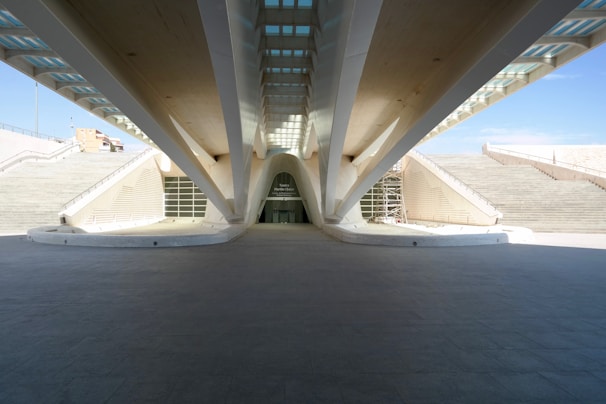 A modern architectural structure with prominent support beams and a symmetrical design. The building is framed by large staircases on either side, leading to an open entrance in the center. Natural light illuminates the area from above, highlighting clean lines and geometric shapes.