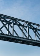 A metal bridge structure with crisscrossing beams spans horizontally against a clear blue sky. The bridge includes a mesh pattern and appears to be part of an industrial or modern architectural design.