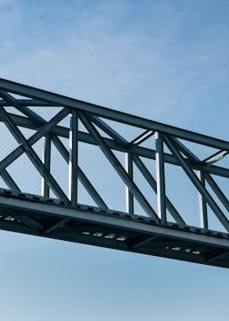 A metal bridge structure with crisscrossing beams spans horizontally against a clear blue sky. The bridge includes a mesh pattern and appears to be part of an industrial or modern architectural design.