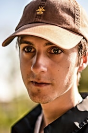 A young man wearing a brown cap with an emblem in the center is looking directly at the viewer. The cap has a casual design and there are visible stubble on his face. The background is blurred, suggesting an outdoor setting.
