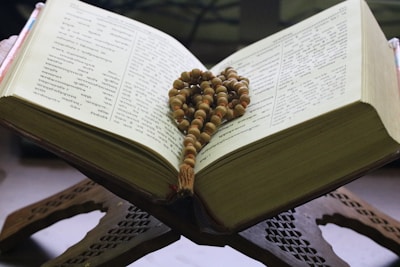 An open book with text in a script resembling Sanskrit, placed on a carved wooden stand. A string of prayer beads is draped across the pages, suggesting a religious context.