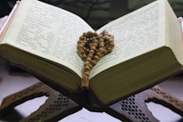 An open book with text in a script resembling Sanskrit, placed on a carved wooden stand. A string of prayer beads is draped across the pages, suggesting a religious context.