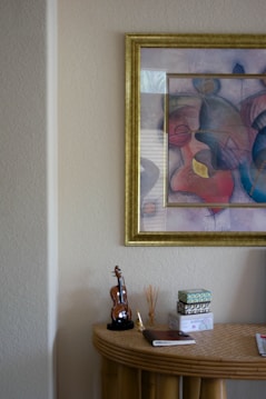 A decorative arrangement featuring a small table topped with a miniature violin, a notepad, a reed diffuser, and a few small boxes. Above the table hangs a colorful abstract painting in a golden frame. The wall is a neutral tone, providing a subtle backdrop to the vibrant art piece.