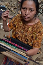 A woman is engaged in weaving, using a traditional backstrap loom. She is wearing a richly patterned brown blouse featuring floral designs. Her focused expression and the intricate setup of threads and wooden rods highlight the artisanal craft.
