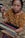 A woman is engaged in weaving, using a traditional backstrap loom. She is wearing a richly patterned brown blouse featuring floral designs. Her focused expression and the intricate setup of threads and wooden rods highlight the artisanal craft.