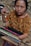 A woman is engaged in weaving, using a traditional backstrap loom. She is wearing a richly patterned brown blouse featuring floral designs. Her focused expression and the intricate setup of threads and wooden rods highlight the artisanal craft.