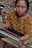 A woman is engaged in weaving, using a traditional backstrap loom. She is wearing a richly patterned brown blouse featuring floral designs. Her focused expression and the intricate setup of threads and wooden rods highlight the artisanal craft.