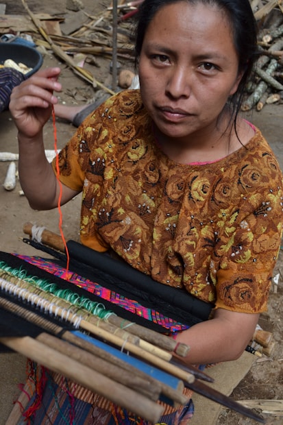 A woman is engaged in weaving, using a traditional backstrap loom. She is wearing a richly patterned brown blouse featuring floral designs. Her focused expression and the intricate setup of threads and wooden rods highlight the artisanal craft.