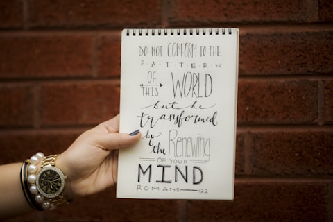 A hand holding a spiral-bound notebook with a quote written in artistic calligraphy. The quote reads, 'Do not conform to the pattern of this world, but be transformed by the renewing of your mind,' attributed to Romans 12:2. The background is a red brick wall, and the hand is adorned with several bracelets, including a watch and a pearl bracelet.