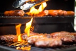 A close-up of a barbecue grill with flames rising from the bottom. On the lower rack, several beef patties are cooking, and on the upper rack, sausages are being grilled. A pair of tongs is being used to handle the food.