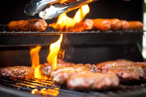 A close-up of a barbecue grill with flames rising from the bottom. On the lower rack, several beef patties are cooking, and on the upper rack, sausages are being grilled. A pair of tongs is being used to handle the food.