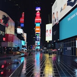 Times Square at night with illuminated billboards and digital advertisements casting colorful reflections on the wet pavement. Few people and taxis are visible, adding to the urban atmosphere.