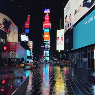 Times Square at night with illuminated billboards and digital advertisements casting colorful reflections on the wet pavement. Few people and taxis are visible, adding to the urban atmosphere.