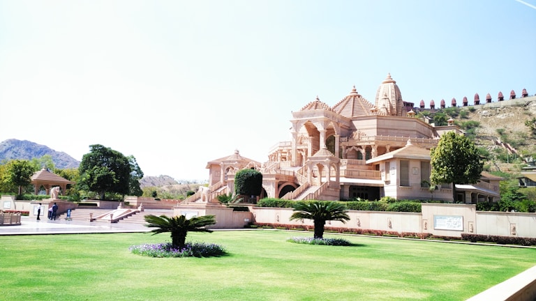 A large, intricately designed temple with detailed carvings stands prominently, surrounded by a manicured green lawn and a few trees. The temple features several domes and arches, and its light pink stone contrasts against the clear blue sky. In the background, there is a hill with small structures along its ridge.