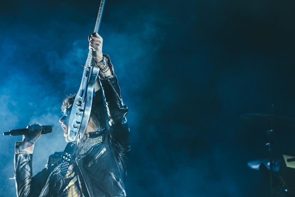 A musician energetically performs on stage, holding a microphone and raising an electric guitar triumphantly. The individual is dressed in a black leather jacket adorned with studs, set against a backdrop of vibrant blue stage lights and smoke, creating a dynamic and dramatic scene.