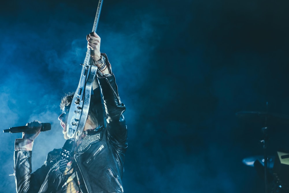 man holding electric guitar and microphone in front of lights inside dim room with smoke