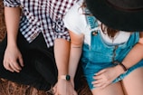 Two people are sitting closely next to each other, holding hands. One is wearing a checked shirt and black pants, while the other is wearing denim overalls and a black hat. A tattoo with the word 'Gratidão' is visible on one arm.