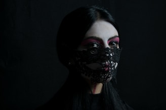 A person with long dark hair is wearing intricately designed black lace covering parts of their face. The use of face paint with a white base and pink accents around the eyes creates a striking contrast against the dark background, adding a mysterious and artistic effect.
