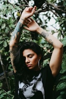 A person with closed eyes stands amidst dense, leafy greenery. Their arms are raised above their head, revealing intricate tattoos on both arms. The individual wears large hoop earrings and a dark shirt with an artistic design.