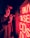 A person wearing a cap is holding a camera and standing next to a bright red neon sign displaying the words 'BUY & SELL CDS'. The neon lights cast a red glow over the individual's face and surroundings, creating a dramatic and moody atmosphere.