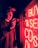A person wearing a cap is holding a camera and standing next to a bright red neon sign displaying the words 'BUY & SELL CDS'. The neon lights cast a red glow over the individual's face and surroundings, creating a dramatic and moody atmosphere.