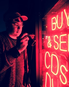 A person wearing a cap is holding a camera and standing next to a bright red neon sign displaying the words 'BUY & SELL CDS'. The neon lights cast a red glow over the individual's face and surroundings, creating a dramatic and moody atmosphere.