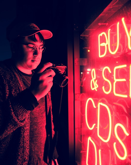 A person wearing a cap is holding a camera and standing next to a bright red neon sign displaying the words 'BUY & SELL CDS'. The neon lights cast a red glow over the individual's face and surroundings, creating a dramatic and moody atmosphere.