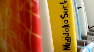 A row of surfboards with various colors and designs, including a yellow one prominently labeled 'Mundaka Surf'. The boards are lined up closely together, highlighting their vibrant hues and patterns.
