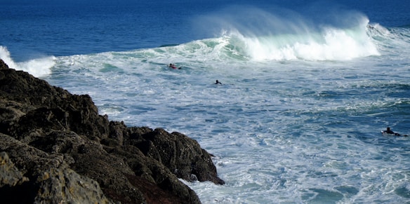 Waves crash against rocky cliffs with surfers venturing out into the ocean, creating a dynamic scene of water and adventure.
