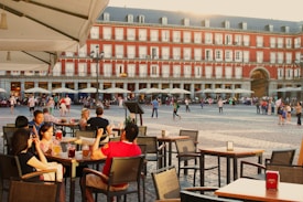 A lively outdoor scene in a historic square with people sitting at cafe tables enjoying drinks, while others walk around the plaza. The background features a large, charming building with a red facade and multiple windows, and a series of umbrellas shading more tables.