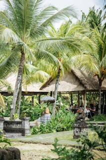 Lush tropical garden with numerous palm trees and vibrant green foliage. Traditional thatched-roof huts blend seamlessly into the natural surroundings, creating a shaded area where people gather. Intricately carved stone statues are nestled among the plants, adding a serene and cultural atmosphere.