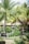 Lush tropical garden with numerous palm trees and vibrant green foliage. Traditional thatched-roof huts blend seamlessly into the natural surroundings, creating a shaded area where people gather. Intricately carved stone statues are nestled among the plants, adding a serene and cultural atmosphere.