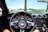 View from the driver's seat of a car, showing the dashboard and steering wheel with a Mustang logo. The scene outside the windshield features a beach setting with palm trees, a pier, and a few people in the distance.