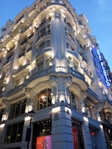 A grand, ornate building with elaborate architectural details, including columns and decorative moldings, is prominently illuminated. The structure has a luxurious appearance, enhanced by strategic lighting that highlights its features. A blue sign with the words 'Hotel Atl&aacute;ntico' is visible on the right side, adding to the opulent atmosphere.
