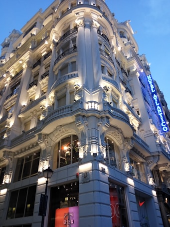 A grand, ornate building with elaborate architectural details, including columns and decorative moldings, is prominently illuminated. The structure has a luxurious appearance, enhanced by strategic lighting that highlights its features. A blue sign with the words 'Hotel Atlántico' is visible on the right side, adding to the opulent atmosphere.