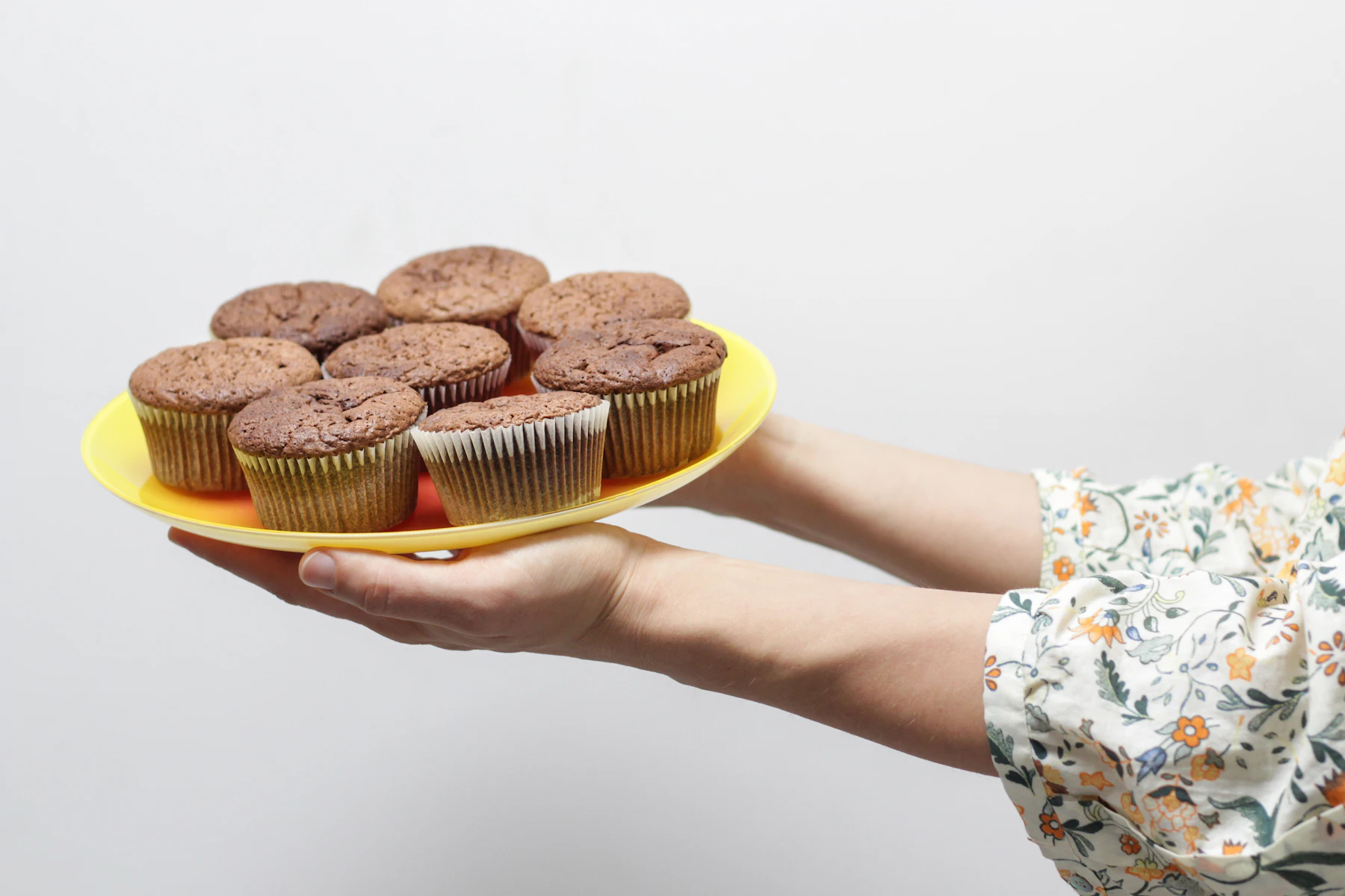 A hand holding out a yellow plate of chocolate muffins in paper liners