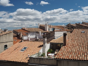 An expanse of terracotta rooftops typical of a Mediterranean village or town is shown under a partly cloudy sky. The roofs have several chimneys and antennas visible, with some small balconies and terraces. There is a table and chairs on one of the terraces, indicating a cozy and lived-in atmosphere.