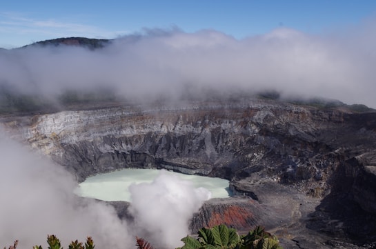 A volcanic crater with a milky turquoise lake is surrounded by rugged, rocky terrain. Clouds hover above, partially obscuring the top rim of the crater. Green foliage is visible in the foreground, adding contrast to the stark landscape.