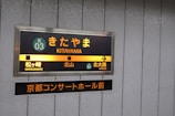 A subway or train station sign features the stop 'Kitayama' with directional indicators pointing towards the next destinations, Matsugasaki to the left and Kitaoji to the right. The sign includes both Japanese characters and their English translations, with a distinct color scheme of orange, yellow, and green. Below, there is an additional sign in Japanese that appears to refer to a concert hall in Kyoto.