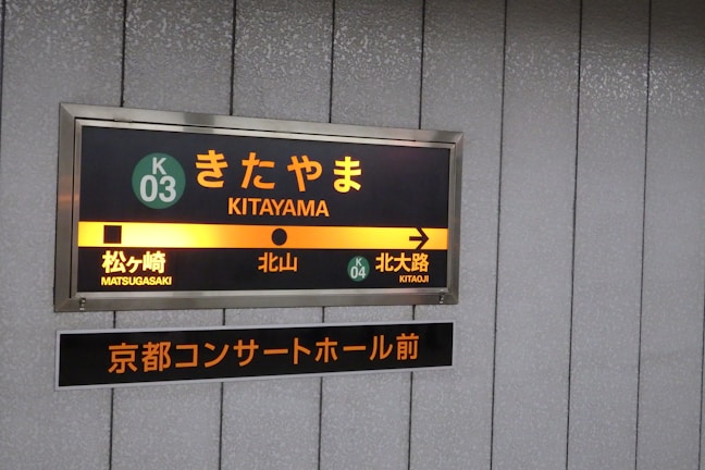 A subway or train station sign features the stop 'Kitayama' with directional indicators pointing towards the next destinations, Matsugasaki to the left and Kitaoji to the right. The sign includes both Japanese characters and their English translations, with a distinct color scheme of orange, yellow, and green. Below, there is an additional sign in Japanese that appears to refer to a concert hall in Kyoto.