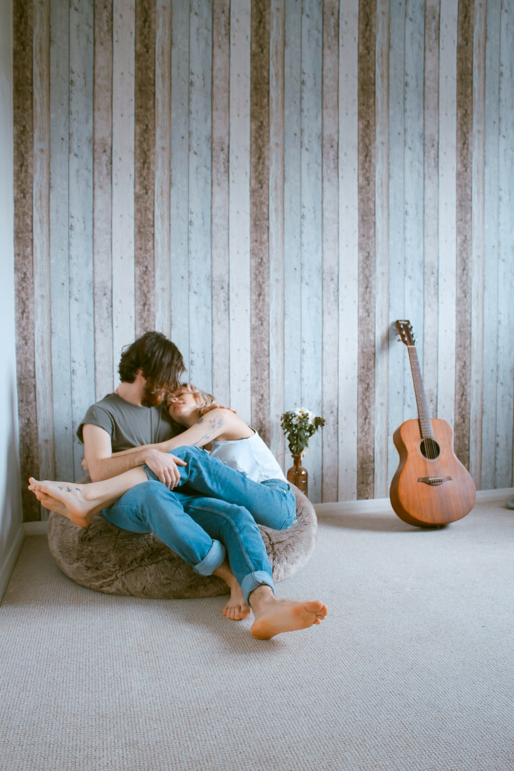 A barefooted man and woman cuddle on a beanbag chair against wood paneling, near a guitar