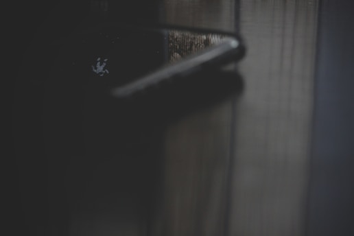 A close-up of a dark-colored door handle with a faint reflection visible on a wooden surface. The image appears to be taken in low light, creating a subtle, mysterious atmosphere.