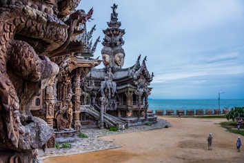 An intricately carved wooden temple featuring ornate sculptures and detailed carvings of deities and mythical creatures. The structure is situated near a sandy area overlooking the sea, with a few people walking nearby.