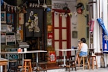 A small, cozy outdoor cafe with a variety of eclectic decorations and posters. A woman is sitting at a table using her phone, with stools surrounding the tables. The cafe entrance is adorned with a traditional fabric print and colorful bunting. The setting suggests an inviting and vibrant atmosphere with a mix of cultural influences.