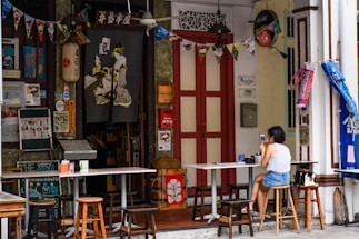 A small, cozy outdoor cafe with a variety of eclectic decorations and posters. A woman is sitting at a table using her phone, with stools surrounding the tables. The cafe entrance is adorned with a traditional fabric print and colorful bunting. The setting suggests an inviting and vibrant atmosphere with a mix of cultural influences.