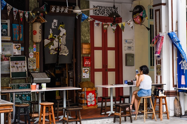 A small, cozy outdoor cafe with a variety of eclectic decorations and posters. A woman is sitting at a table using her phone, with stools surrounding the tables. The cafe entrance is adorned with a traditional fabric print and colorful bunting. The setting suggests an inviting and vibrant atmosphere with a mix of cultural influences.
