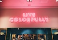 Bright pink neon sign displaying the words 'Live Colorfully' against a light-colored wall. Below the sign, a store-like display with shelves and mannequins can be seen.