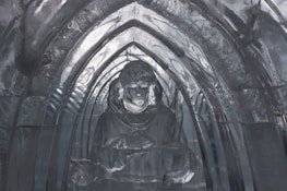 A detailed ice sculpture of a robed figure is captured within an arched setting, with smooth, translucent ice defining the intricate shapes and contours. The arch frames the figure, adding depth and highlighting the skilled craftsmanship in the ice carving.